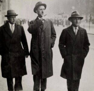 Drei Männer mit Hüten und Mänteln gekleidet. Sie laufen eine Straße entlang und unterhalten sich dabei.